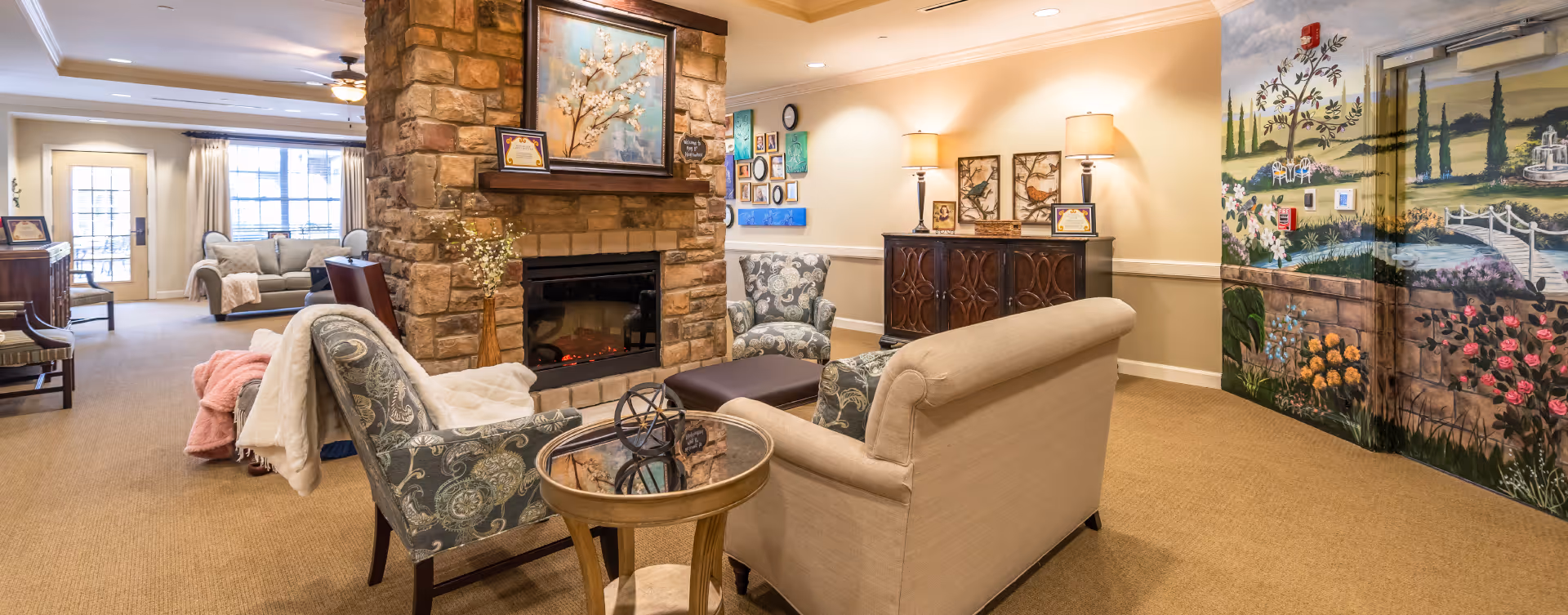 A cozy living room area in a senior living facility featuring a stone fireplace with a painting above it, surrounded by comfortable upholstered chairs and a beige sofa. The room has warm lighting, a decorative side table, framed artwork on the walls, and a colorful mural depicting a garden scene on one wall. Large windows and glass doors allow natural light to fill the space.