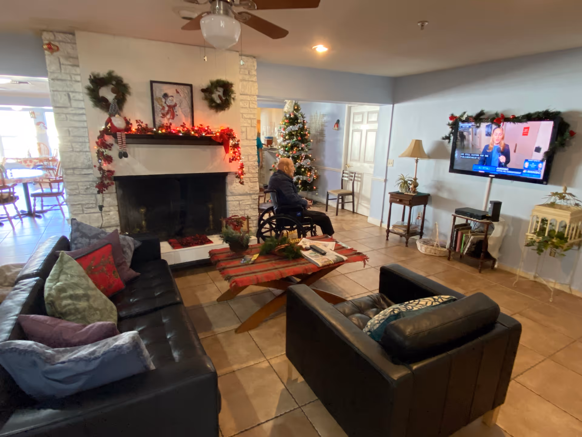 A cozy living room decorated for Christmas with a white stone fireplace adorned with wreaths and red garlands. A Christmas tree stands in the corner near a man sitting in a wheelchair facing a wall-mounted TV. The room has a black leather sofa and chair with colorful pillows, a wooden coffee table with a red plaid cloth, and tile flooring.