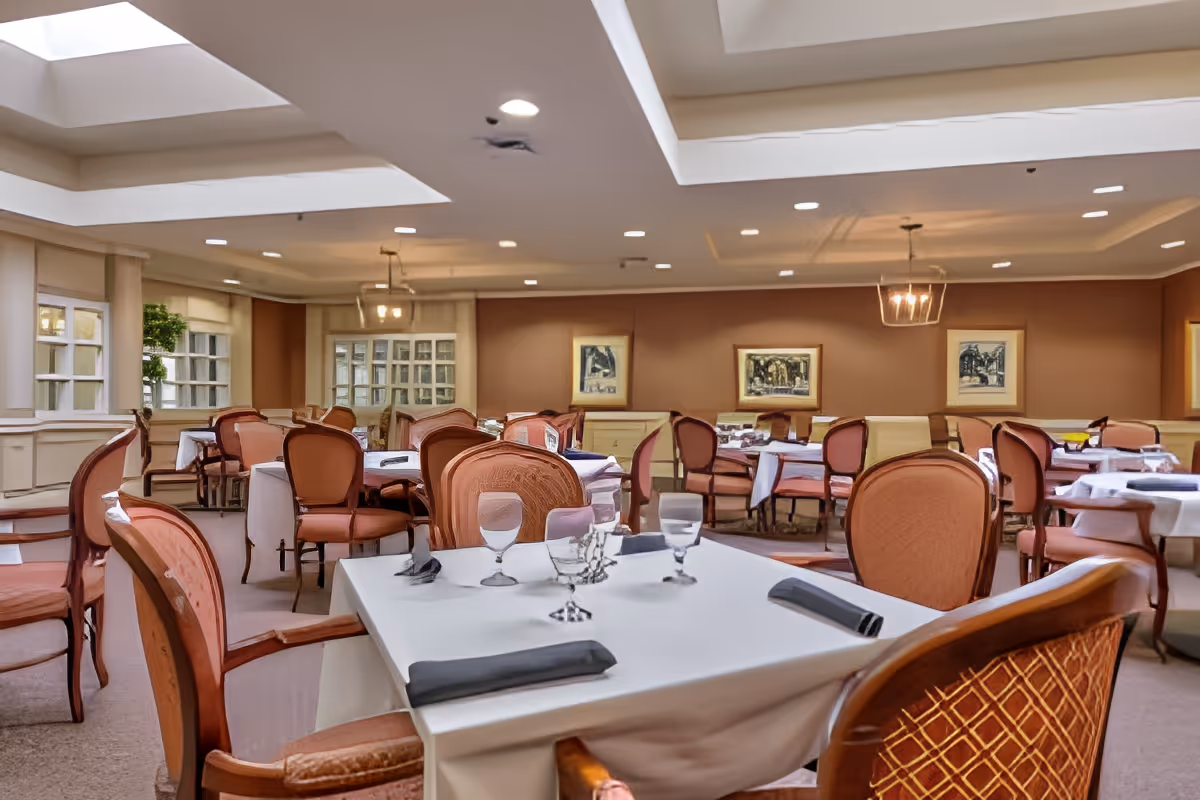 Dining room with neatly set tables, upholstered chairs, artwork on the walls, and recessed ceiling lighting.