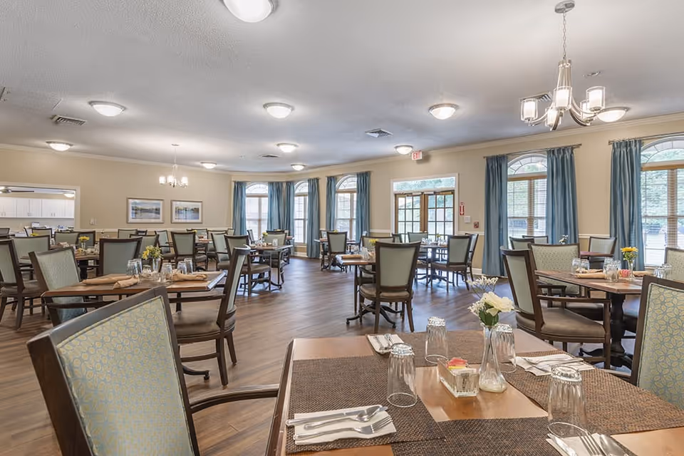 Spacious dining room with multiple set tables and chairs, chandeliers, and large windows with blue curtains.