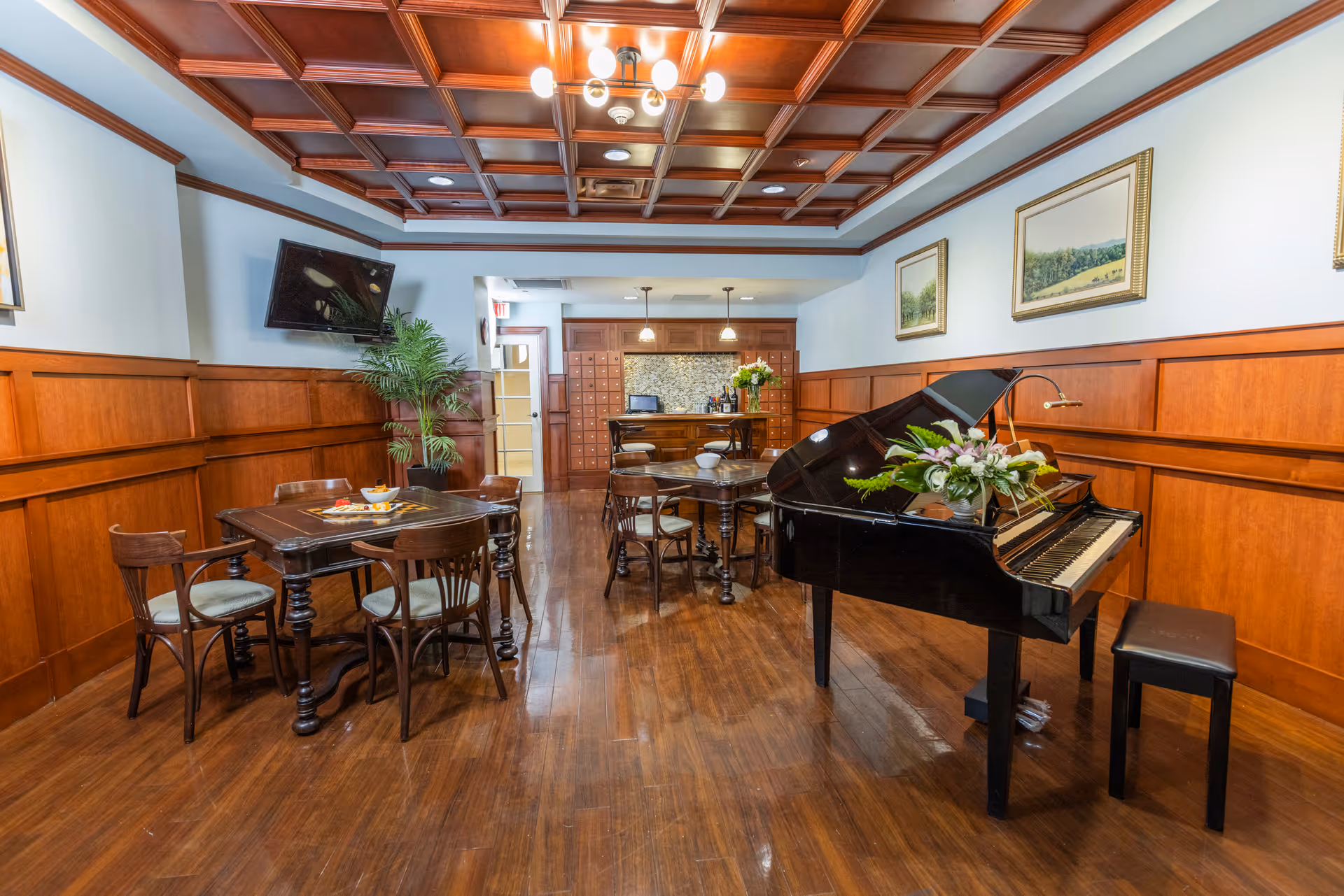 A cozy interior room with wooden paneled walls and a coffered wooden ceiling. The room features a black grand piano with a floral arrangement on top, several wooden tables with chairs, a wall-mounted TV, framed landscape paintings, and a small bar area with pendant lights in the background.