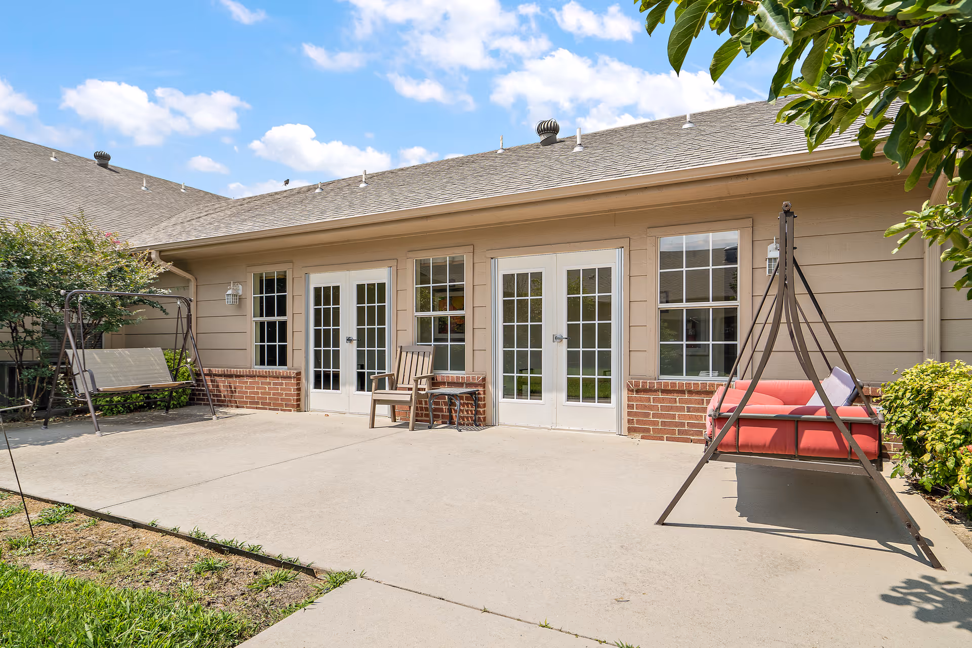 Outdoor patio area with two swing seats, one beige and one red, positioned on a concrete surface in front of a beige building with brick accents and multiple glass-paneled doors and windows under a partly cloudy blue sky.