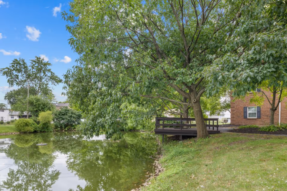 A peaceful outdoor scene at Oakleaf Village of Columbus featuring a small pond with reflections of surrounding trees, a wooden deck extending from the grassy bank, and part of a brick building with windows and black shutters under a blue sky with scattered clouds.