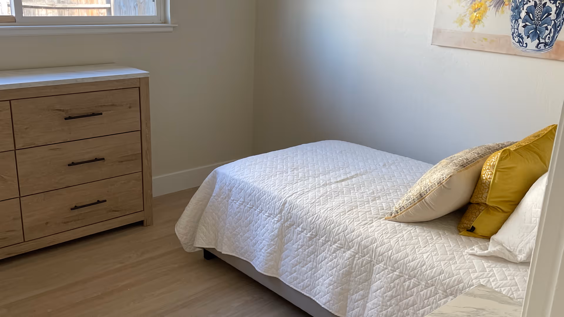 A small bedroom with a single bed covered in a white quilted bedspread and three decorative pillows in shades of yellow and beige. Next to the bed is a wooden dresser with six drawers and black handles. A window above the dresser lets in natural light, and a framed floral artwork hangs on the wall above the bed.
