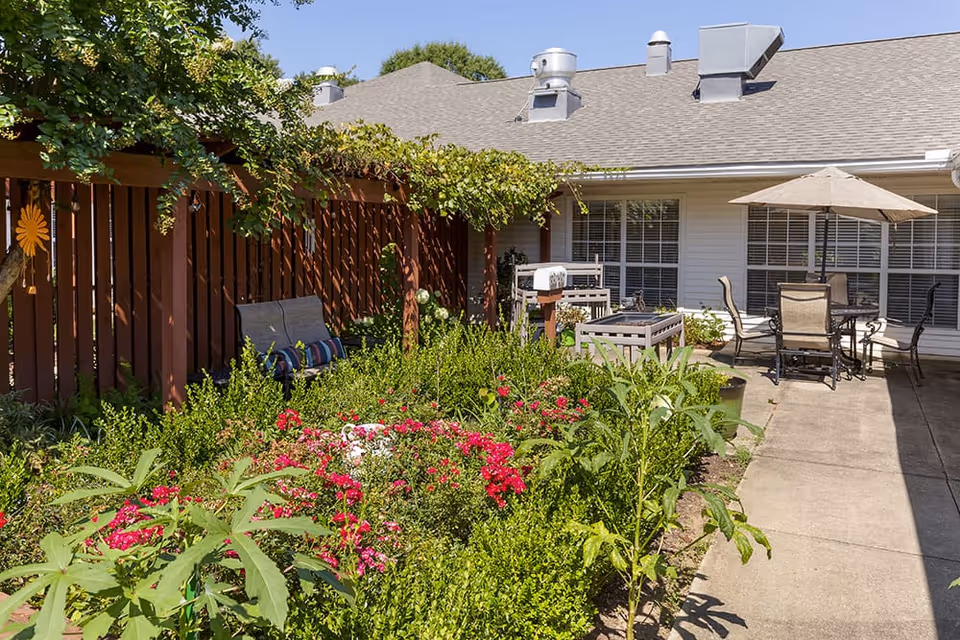 Sunlit courtyard with a flowerbed, wooden pergola, outdoor seating and umbrella beside a single-story building.