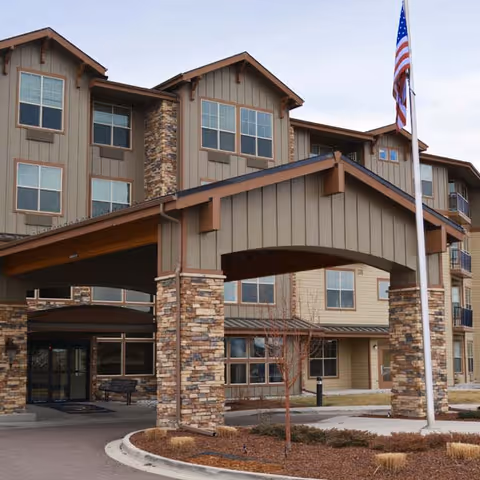 Exterior view of a multi-story senior living facility with stone and wood siding, a covered entrance supported by stone pillars, several windows, and an American flag on a flagpole in front.
