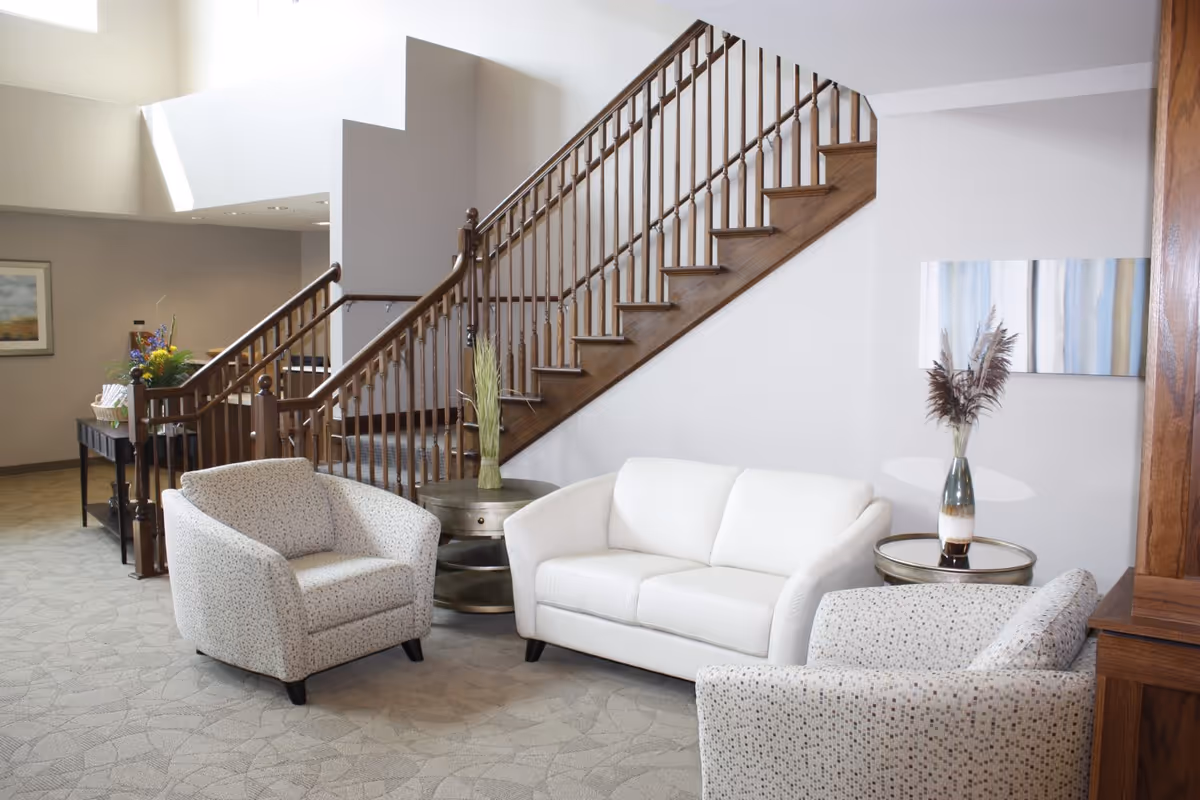 A cozy seating area in a senior living facility featuring a white loveseat and two patterned armchairs arranged around two round side tables with decorative vases. Behind the seating area is a wooden staircase with a railing, and the walls are painted light gray with a piece of abstract art hanging on one side.