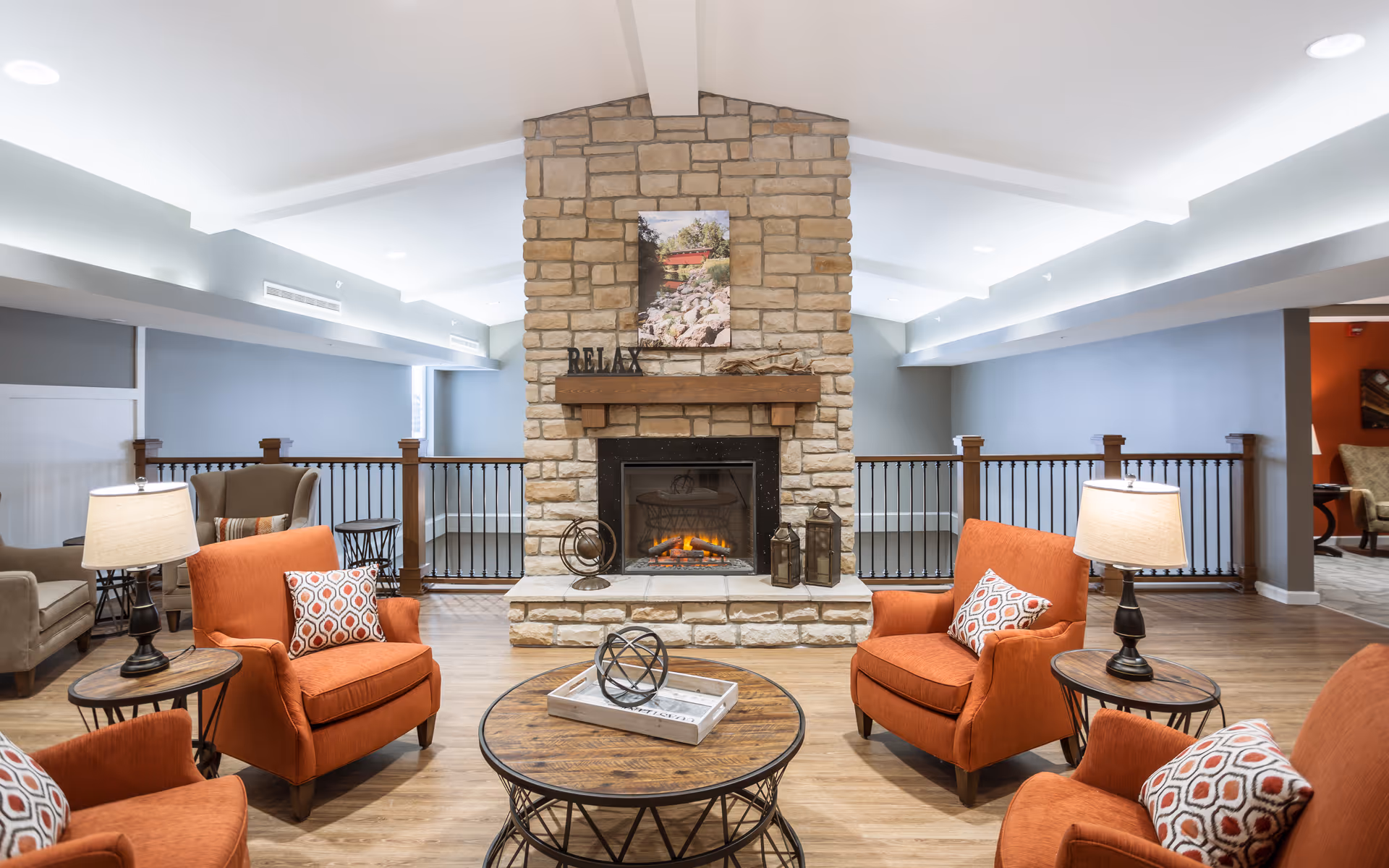 A cozy living room area with a stone fireplace in the center. Four orange armchairs with patterned cushions are arranged around a round wooden coffee table with decorative items. Two side tables with lamps flank the chairs. The room has wooden flooring, light gray walls, and a vaulted ceiling with recessed lighting. A picture hangs above the fireplace with the word RELAX displayed on the mantel.