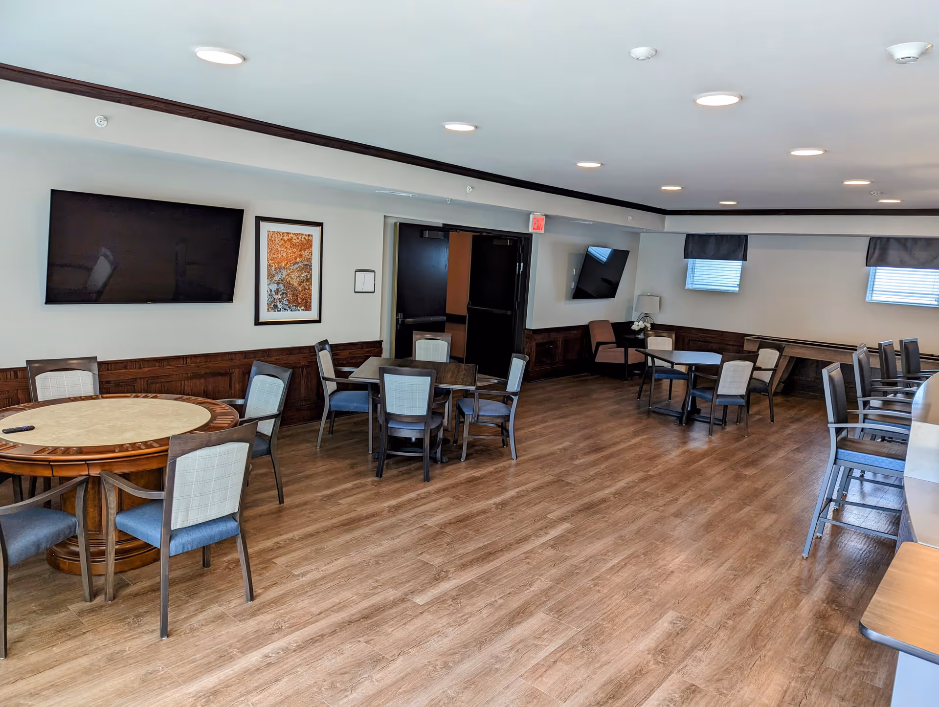 Spacious common dining room with several round and square tables, chairs, wall-mounted TVs, and wood-look flooring.