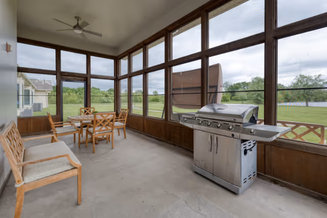 A screened-in porch area with a stainless steel gas grill on the right side and a wooden dining table with four chairs in the center. There is a wooden bench with cushions on the left side. Large windows provide a view of a grassy outdoor area with trees and a pond in the distance.