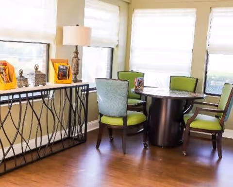 Small dining nook with a round dark table surrounded by green-upholstered chairs, a console table with a lamp and framed photos, and large windows with shades.