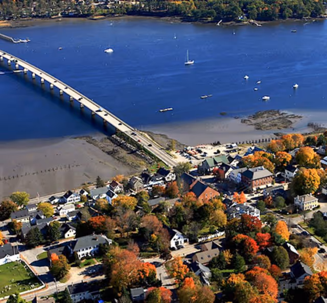 Aerial view of a coastal town with a bridge crossing blue water, sailboats, and houses surrounded by colorful autumn foliage.