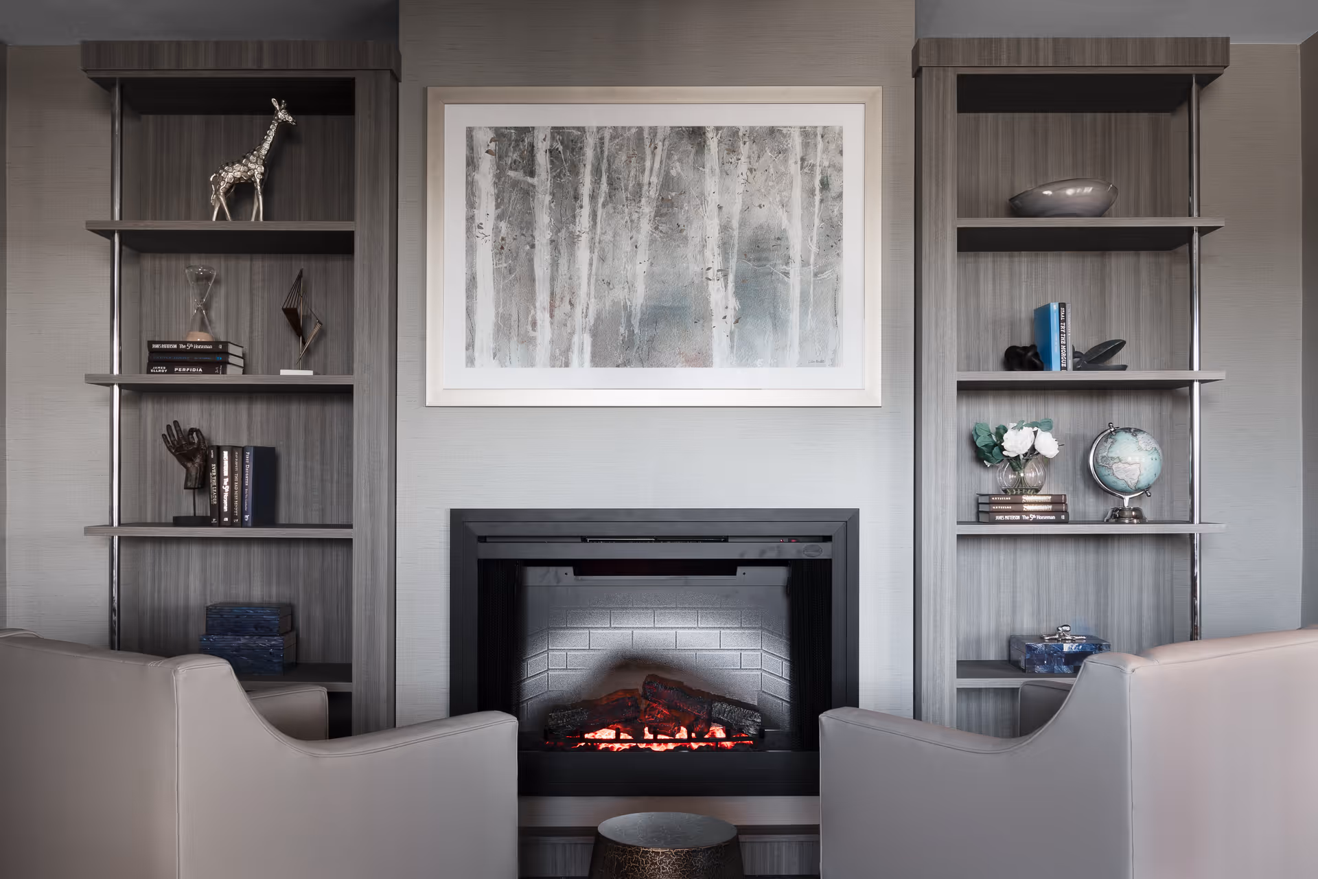 A cozy interior living area featuring a modern electric fireplace centered between two built-in wooden shelves. The shelves are decorated with books, a globe, vases, and small sculptures. Above the fireplace hangs a framed abstract painting with muted colors. Two light gray armchairs face the fireplace with a small round table between them.