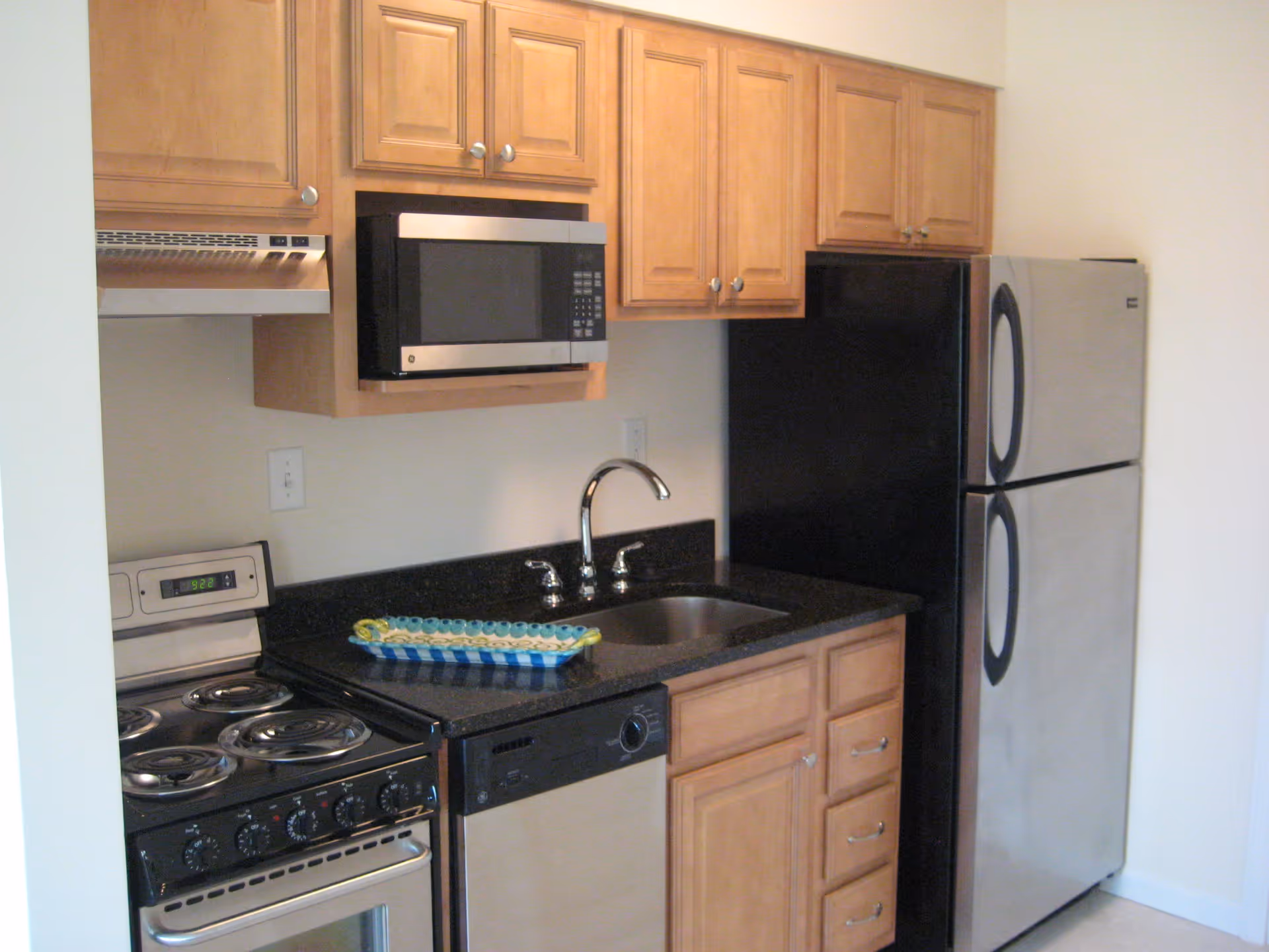 A compact kitchen featuring wooden cabinets, a stainless steel microwave mounted above a black electric stove with four coil burners, a black dishwasher, a stainless steel sink with a chrome faucet, and a stainless steel refrigerator with black trim.