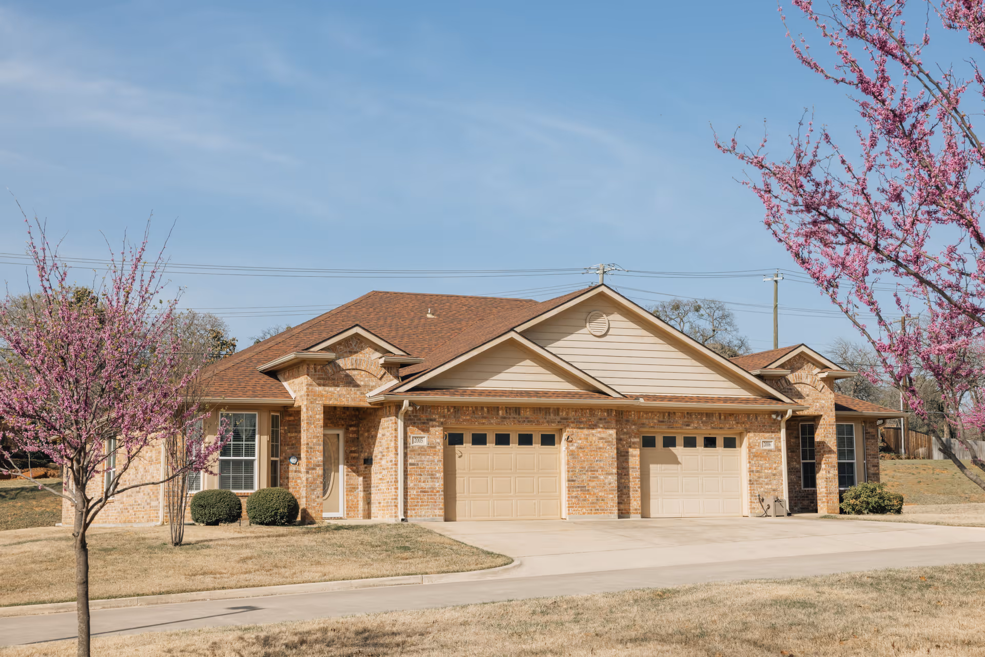 Exterior view of a single-story brick residential building with two garages and a brown shingled roof, surrounded by a lawn and two blossoming trees with pink flowers under a clear blue sky.