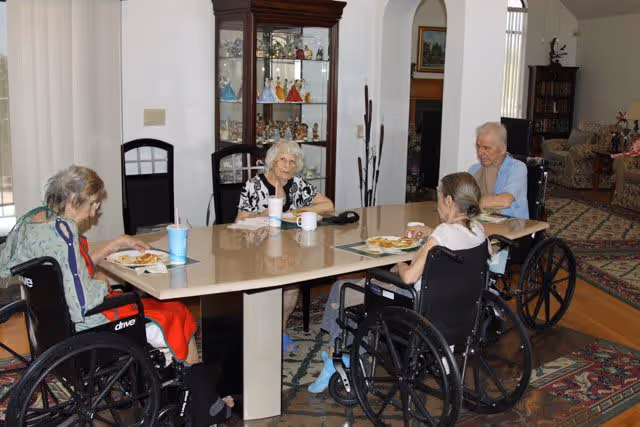 Four elderly individuals, three in wheelchairs, sitting around a rectangular dining table eating a meal in a well-lit room with a display cabinet and living room furniture in the background.