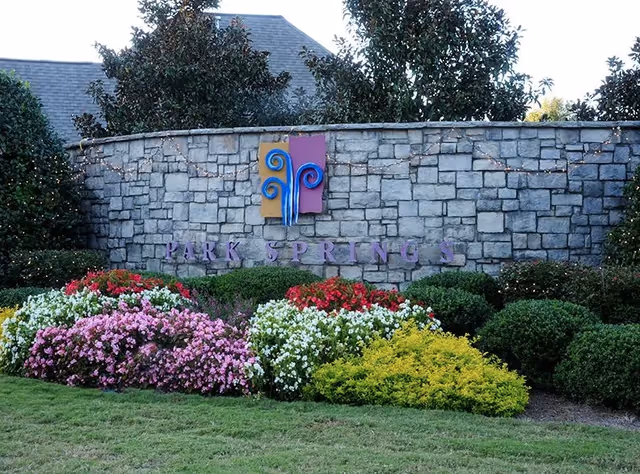 Stone wall with a colorful decorative sign and the words 'PARK SPRINGS' mounted on it, surrounded by neatly trimmed bushes and vibrant flower beds with pink, white, red, and yellow flowers in front, with trees and a building roof visible in the background.