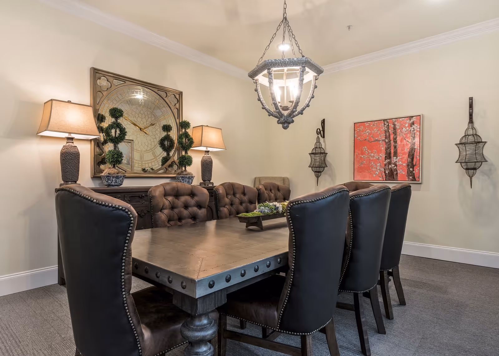 A dining room with a large rectangular wooden table surrounded by eight upholstered chairs, two table lamps on a sideboard, a large decorative clock on the wall, two topiary plants, a hanging chandelier, wall sconces, and a red floral painting on the wall.