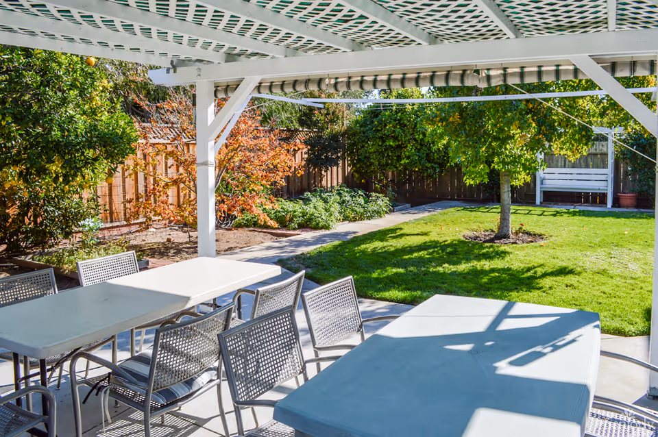 Outdoor patio area with white lattice roof covering, featuring two white tables and several metal chairs with cushions. The patio overlooks a green lawn with a tree, surrounded by a wooden fence and various shrubs and plants.