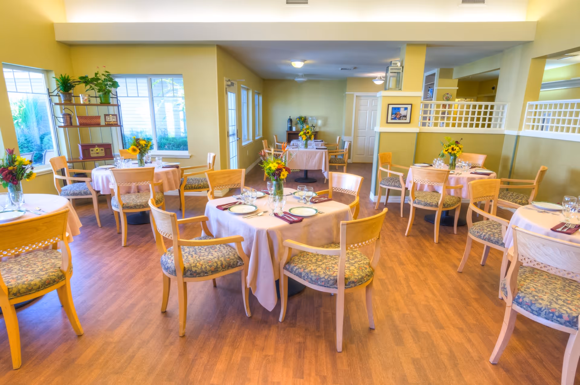 A bright dining room with several round tables covered with light pink tablecloths, each set with plates, glasses, and silverware. Wooden chairs with patterned cushions surround the tables. Vases with colorful flowers are placed on each table. Large windows let in natural light, and there are plants on a metal shelf near the windows. The room has light yellow walls and wood flooring.