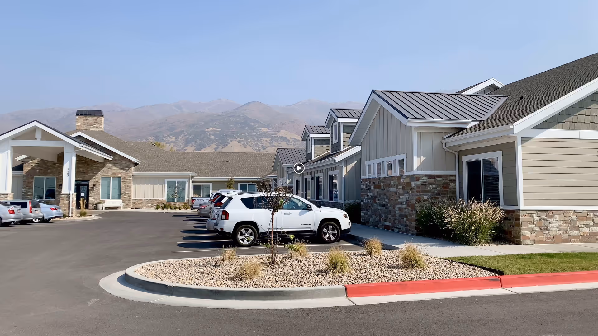 Exterior view of Whisper Cove Assisted Living and Memory Care facility showing a single-story building with stone and siding facade, several parked cars, a landscaped area with rocks and plants, and mountains in the background under a clear sky.