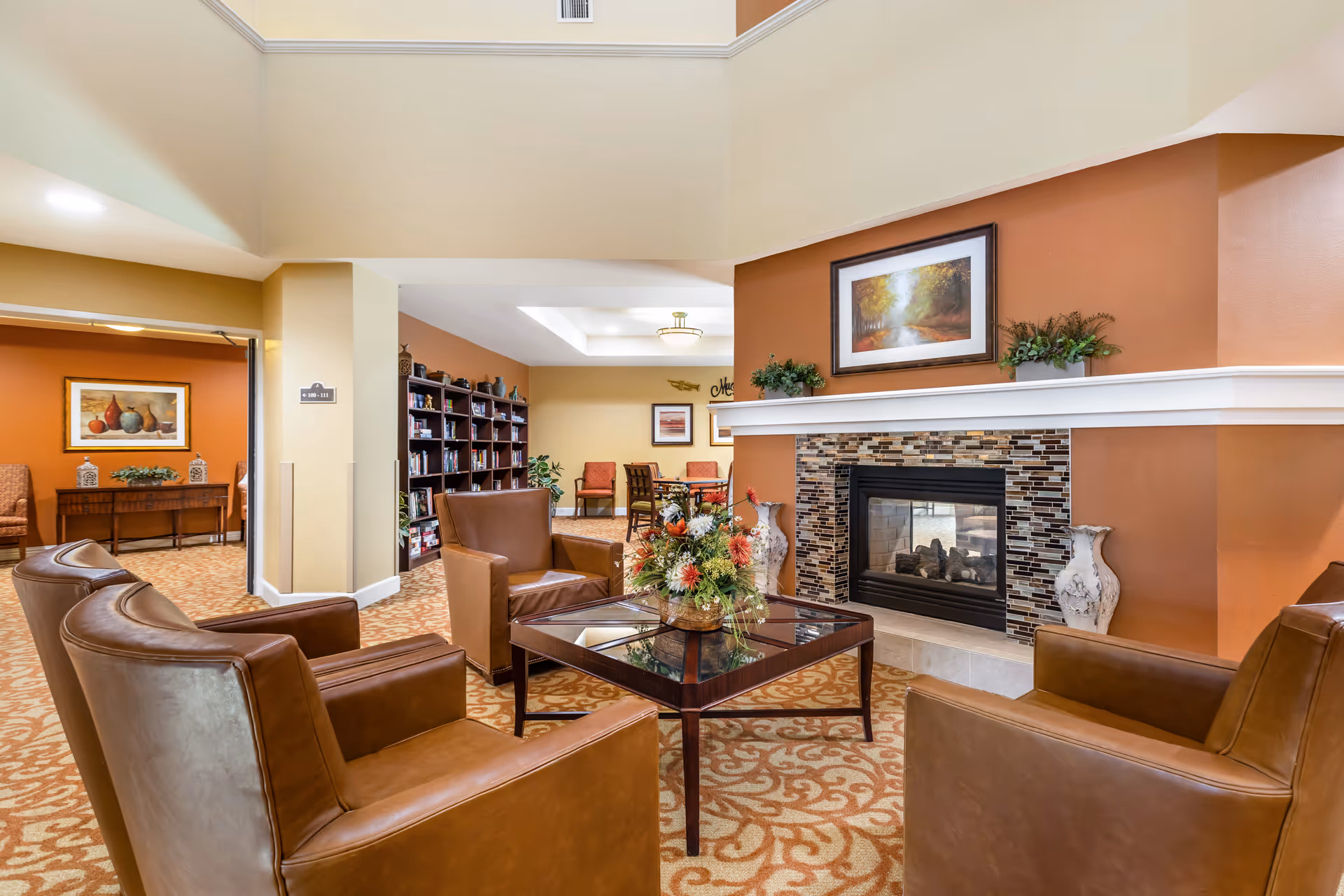 A cozy senior living facility common area with four brown leather armchairs arranged around a glass-top wooden coffee table with a floral centerpiece. The room features a fireplace with a mosaic tile surround, a framed landscape painting above it, and two decorative vases on either side. In the background, there is a bookshelf filled with books and a seating area with chairs and framed artwork on the walls. The space has warm earth-tone colors and patterned carpet.