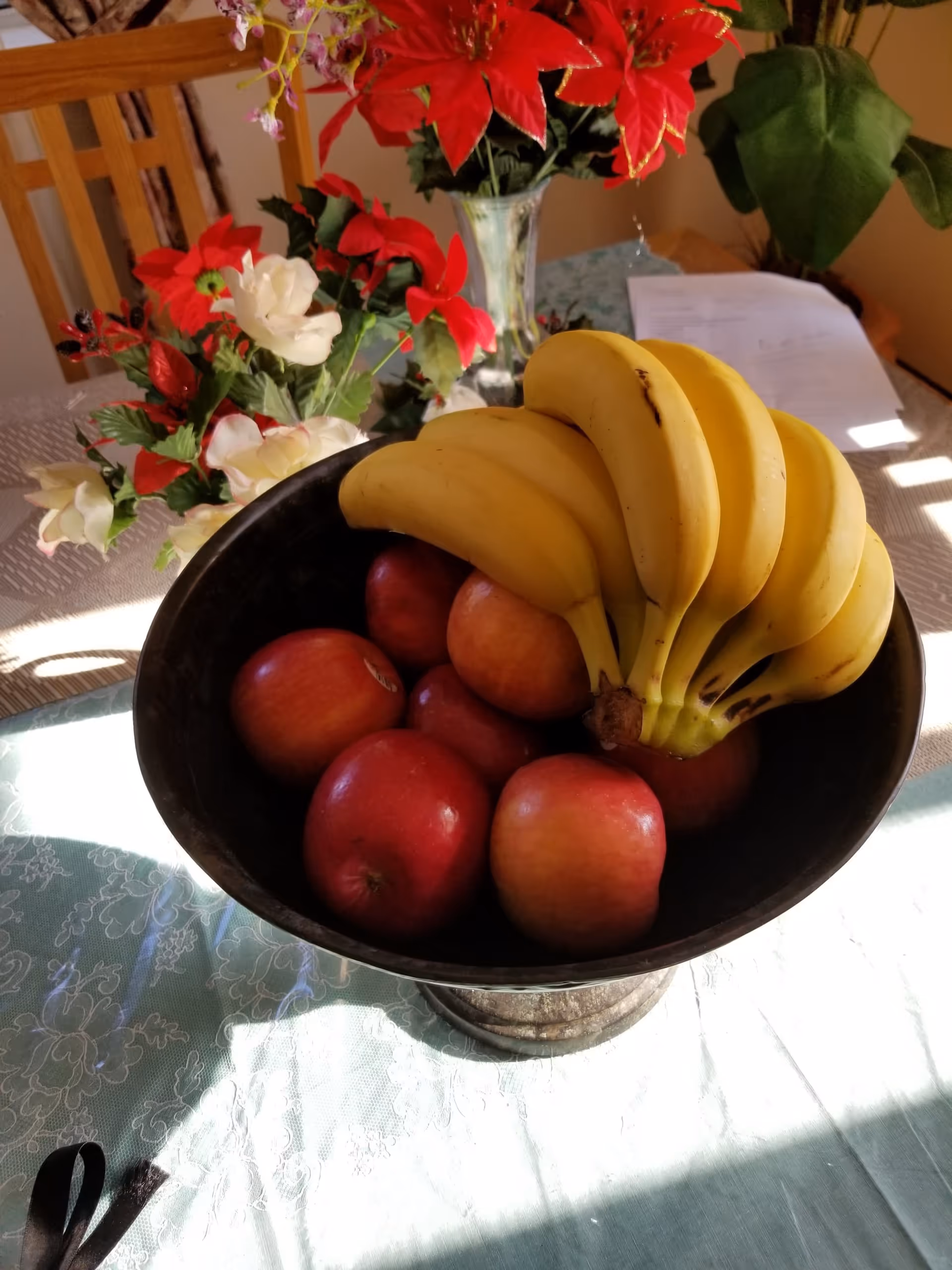 A black bowl filled with red apples and a bunch of bananas on a table covered with a light green tablecloth. Behind the bowl, there is a vase with red and white flowers and a wooden chair. Some papers are also visible on the table in the background.