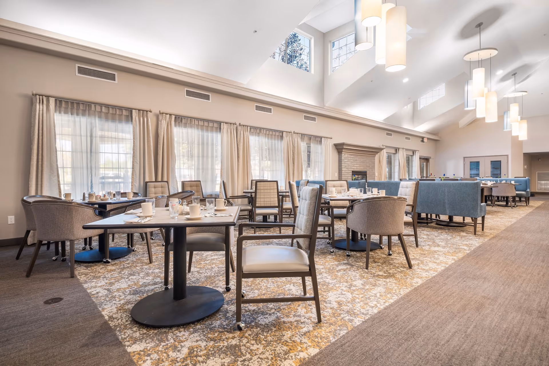 A spacious dining room with multiple tables and chairs arranged neatly. The room features large windows with sheer curtains allowing natural light to fill the space. There are modern pendant lights hanging from a high ceiling with skylights. The tables are set with cups, glasses, and napkins, and there is a fireplace along one wall.