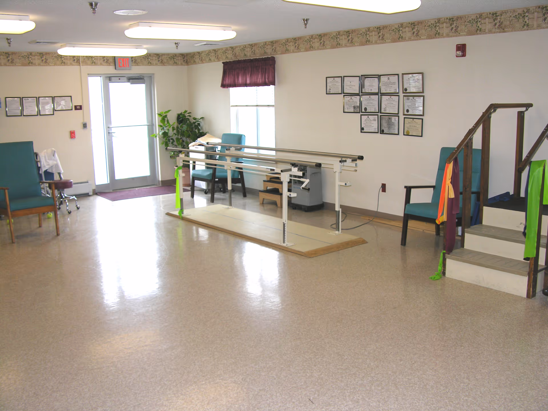 A rehabilitation or physical therapy room with parallel bars for walking exercises, two chairs, a small set of stairs with handrails, certificates on the wall, a window with a purple valance, and an exit door in the background.
