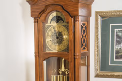 A close-up view of a wooden grandfather clock with a decorative clock face and pendulum, positioned next to a framed picture on a light-colored wall.