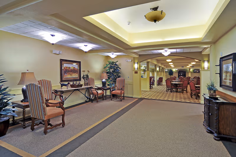 Well-lit interior of a senior living facility with upholstered chairs and decorative tables leading into a dining area with multiple tables and chairs.