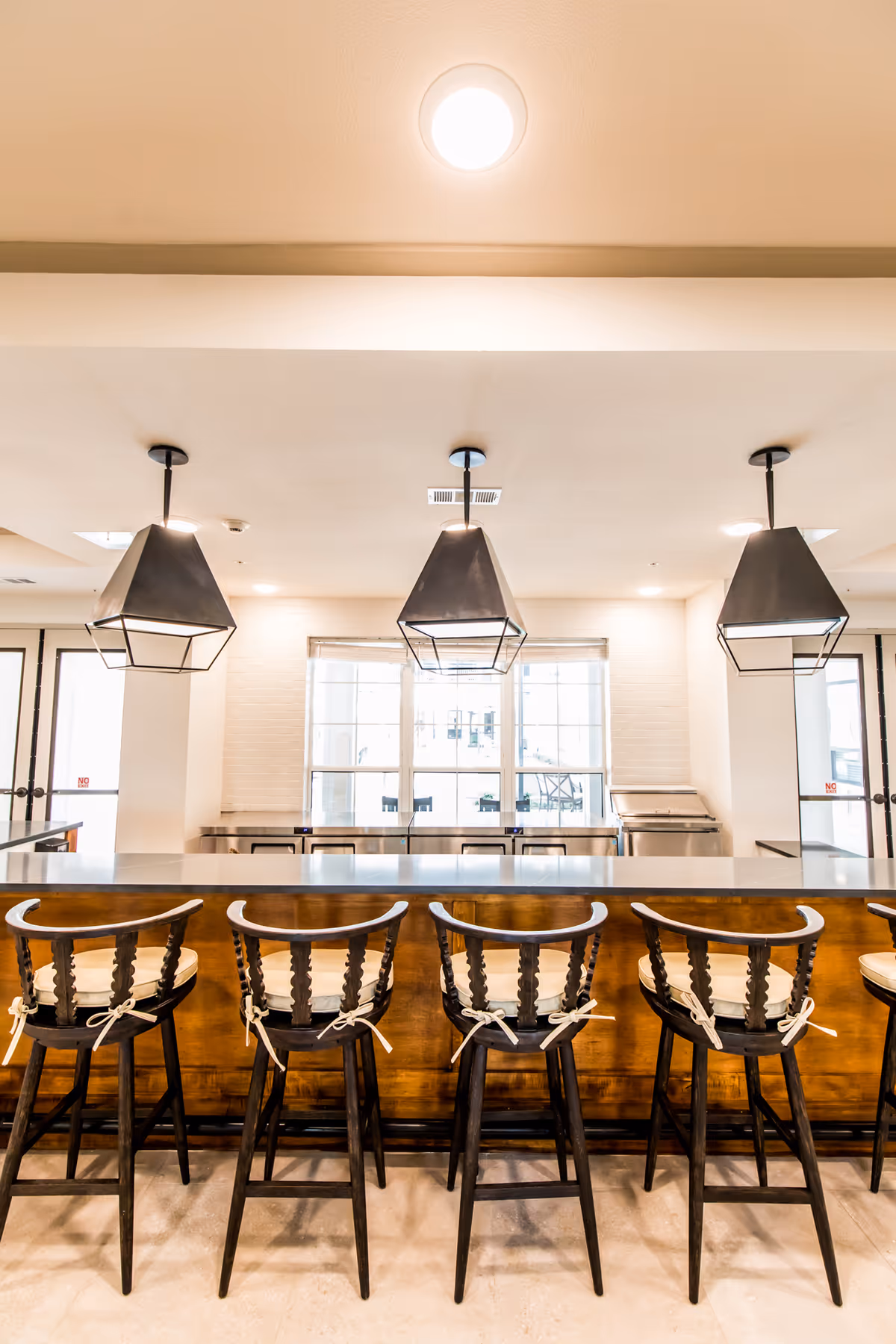 Interior view of a modern kitchen area with a long wooden counter and five high-back bar stools with cushions tied to the seats. Above the counter, there are three black geometric pendant lights hanging from the ceiling. Large windows in the background allow natural light to fill the space.