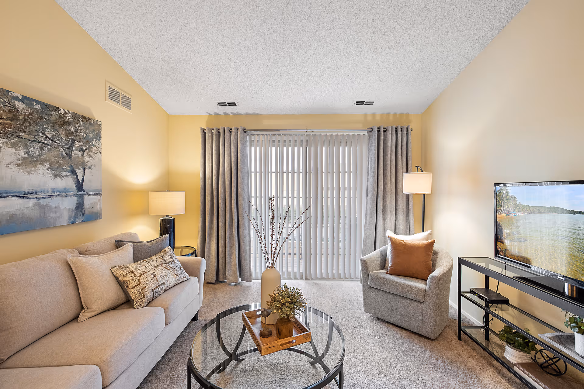 A cozy living room with a beige sofa adorned with multiple pillows on the left, a gray armchair with a brown pillow on the right, and a glass coffee table with decorative items in the center. Behind the armchair is a floor lamp, and behind the sofa is a table lamp. The back wall features large vertical blinds covering a sliding glass door, flanked by gray curtains. A flat-screen TV on a black metal stand is on the right side of the room. The walls are painted light yellow, and a large painting of a tree over water hangs above the sofa.