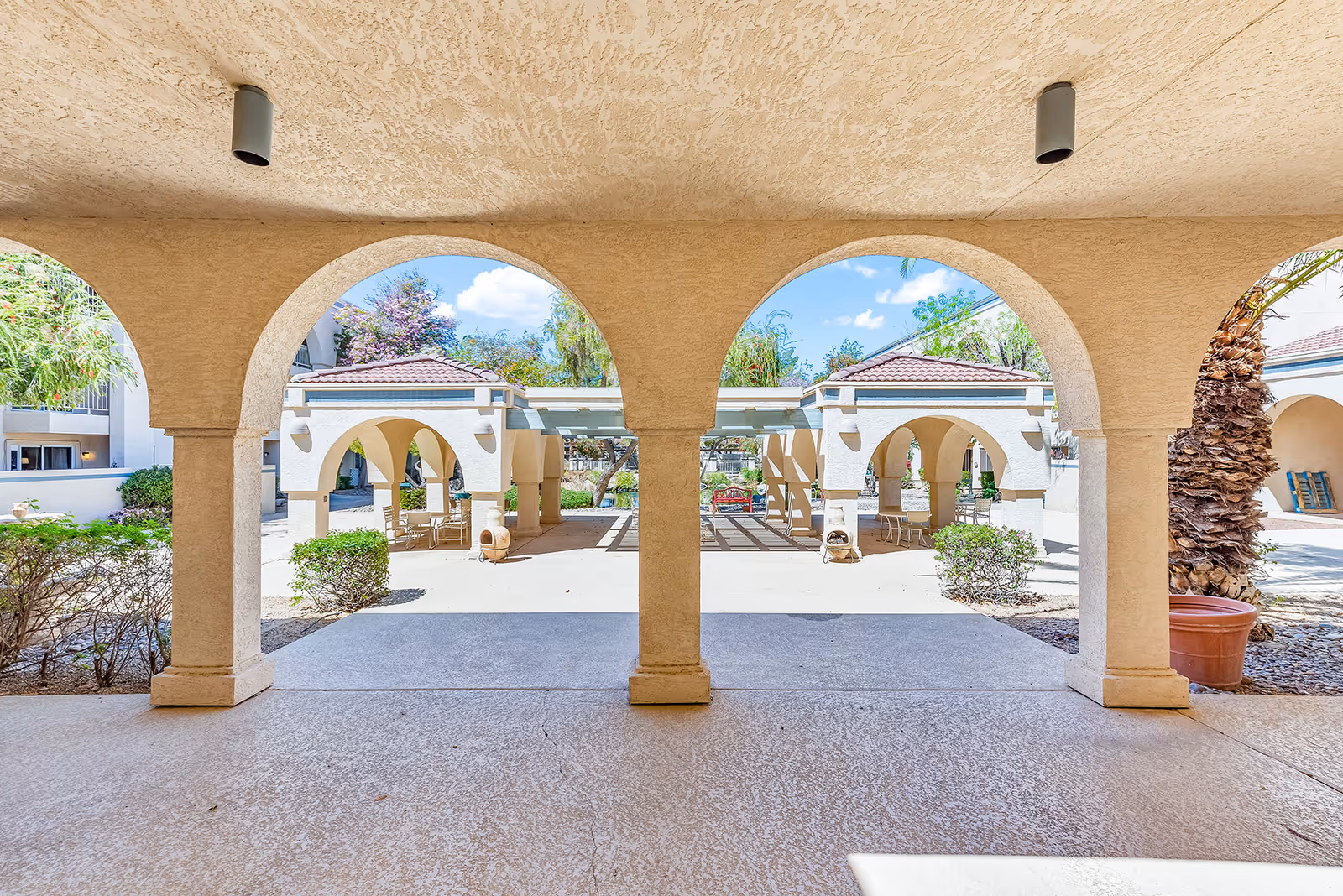 Covered archway framing a sunny outdoor courtyard with tiled roofs, seating, and landscaping at a senior living community.