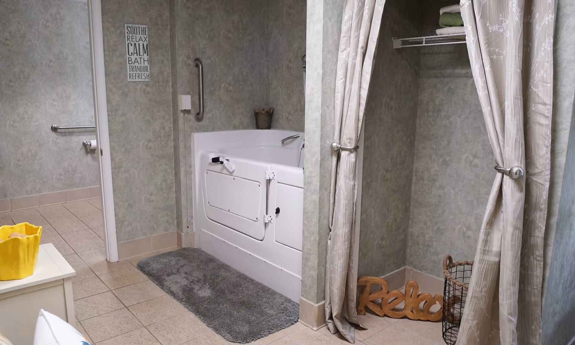 A bathroom with a walk-in bathtub featuring safety handles and a gray bath mat in front. To the right, there is a curtained closet area with towels on a shelf and a decorative wooden sign that says 'Relax'. On the wall above the bathtub, there is a sign with calming words like 'Soothe', 'Relax', 'Calm', 'Bath', 'Tranquil', and 'Refresh'. The floor is tiled and the walls have a textured greenish-gray wallpaper.