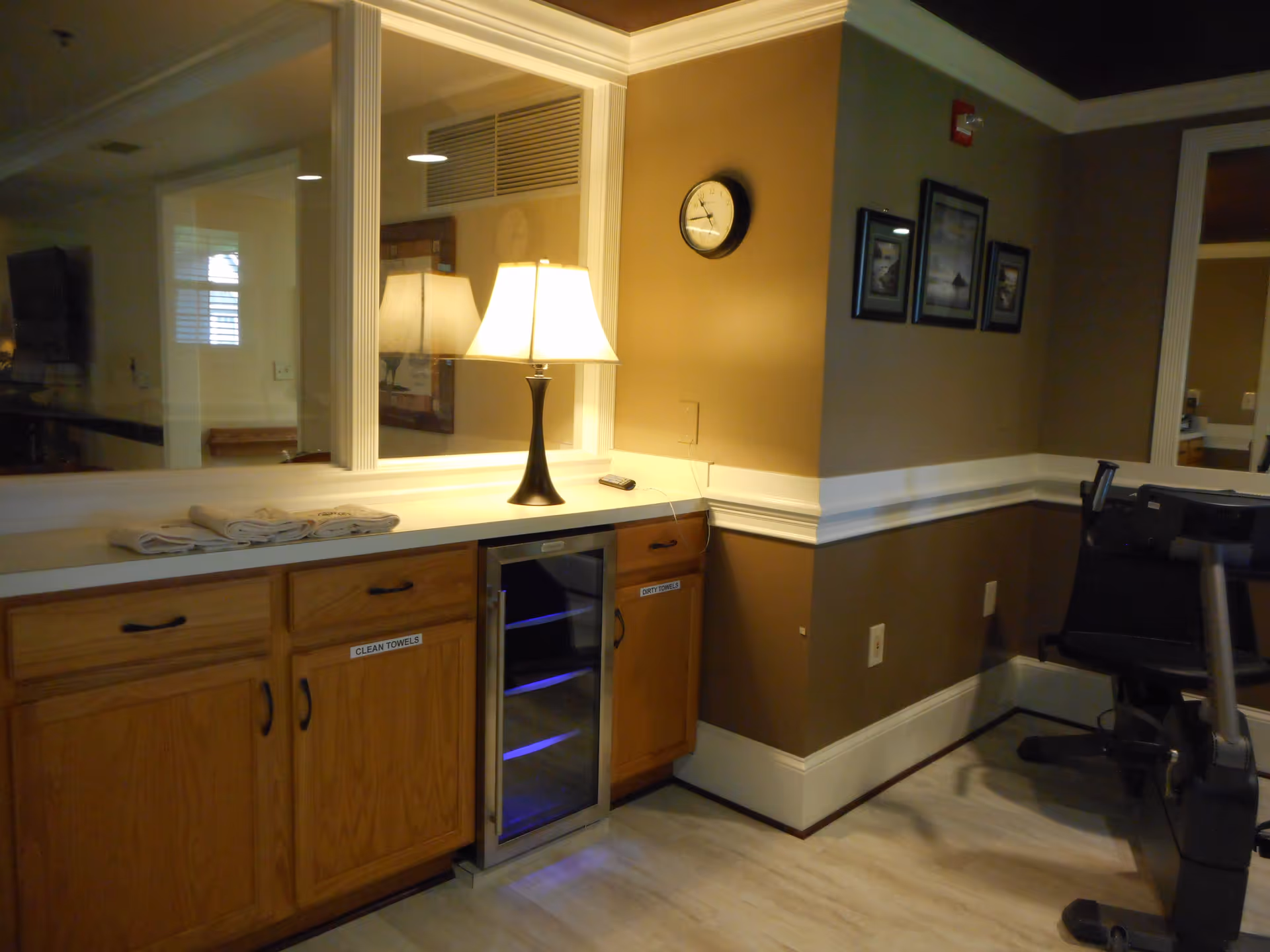Interior view of a room with a wooden cabinet labeled 'CLEAN TOWELS' and 'DIRTY TOWELS', a lamp on the countertop, a wall clock, framed pictures on the wall, and exercise equipment partially visible on the right side.