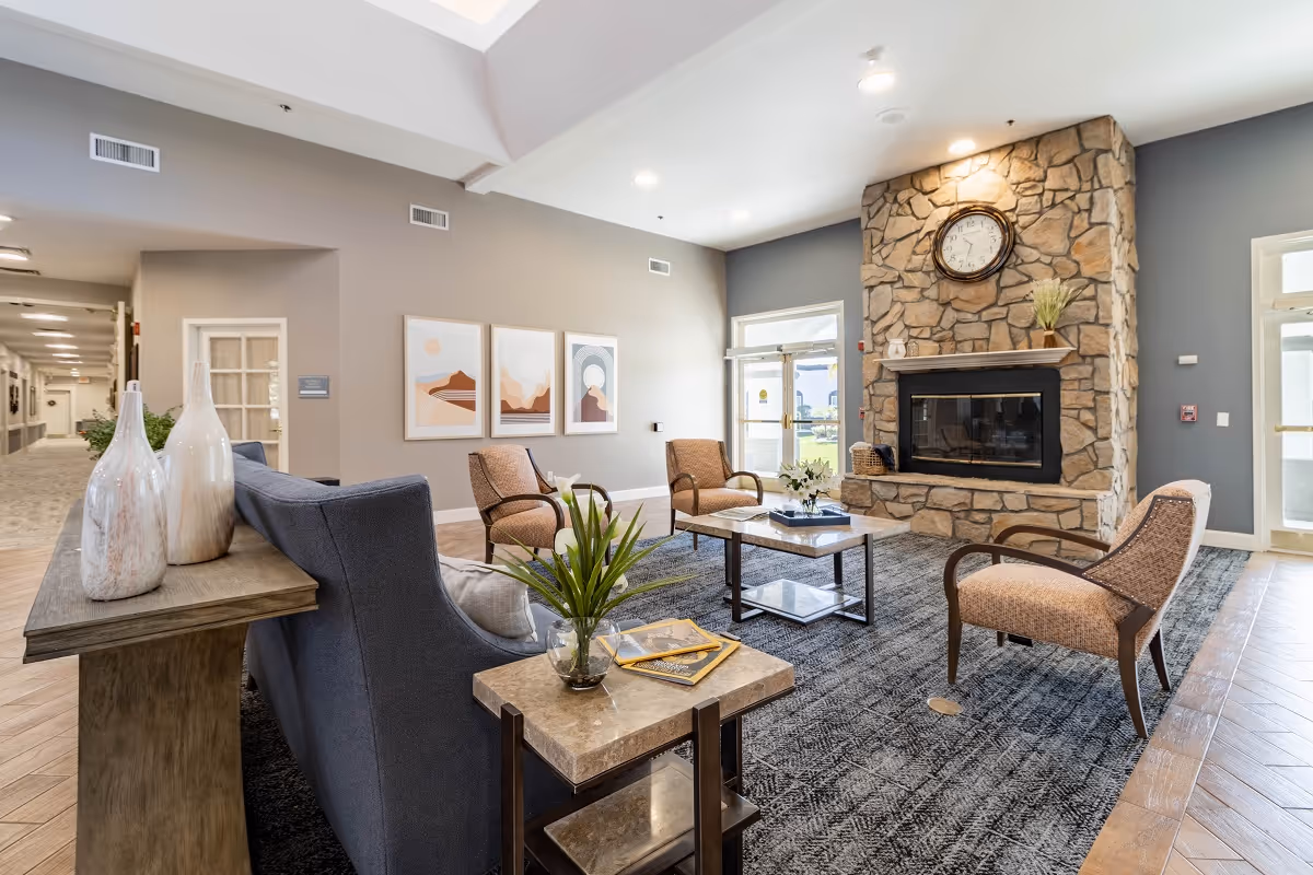 Spacious senior living common area with sofas and chairs arranged around a coffee table and a stone fireplace topped by a wall clock.