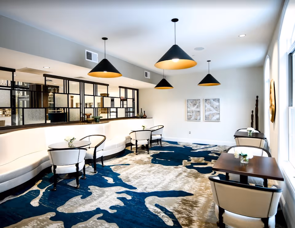 A modern, well-lit dining area with a long curved white bench along one wall, paired with small round tables and cushioned chairs. The floor features a large blue and beige patterned carpet. Four black pendant lights hang from the ceiling. The room has large windows on one side and decorative wall art on the opposite wall.
