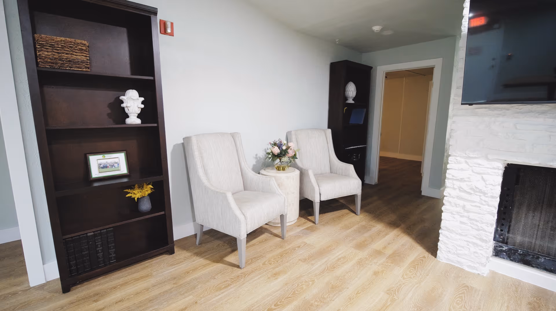 A cozy interior corner of a senior living facility featuring two beige upholstered armchairs with a small round table between them holding a floral arrangement. To the left is a tall dark wooden bookshelf with decorative items and books. On the right side, there is a white stone fireplace with a mounted flat-screen TV above it. The floor is light wood, and there is a doorway leading to another room in the background.
