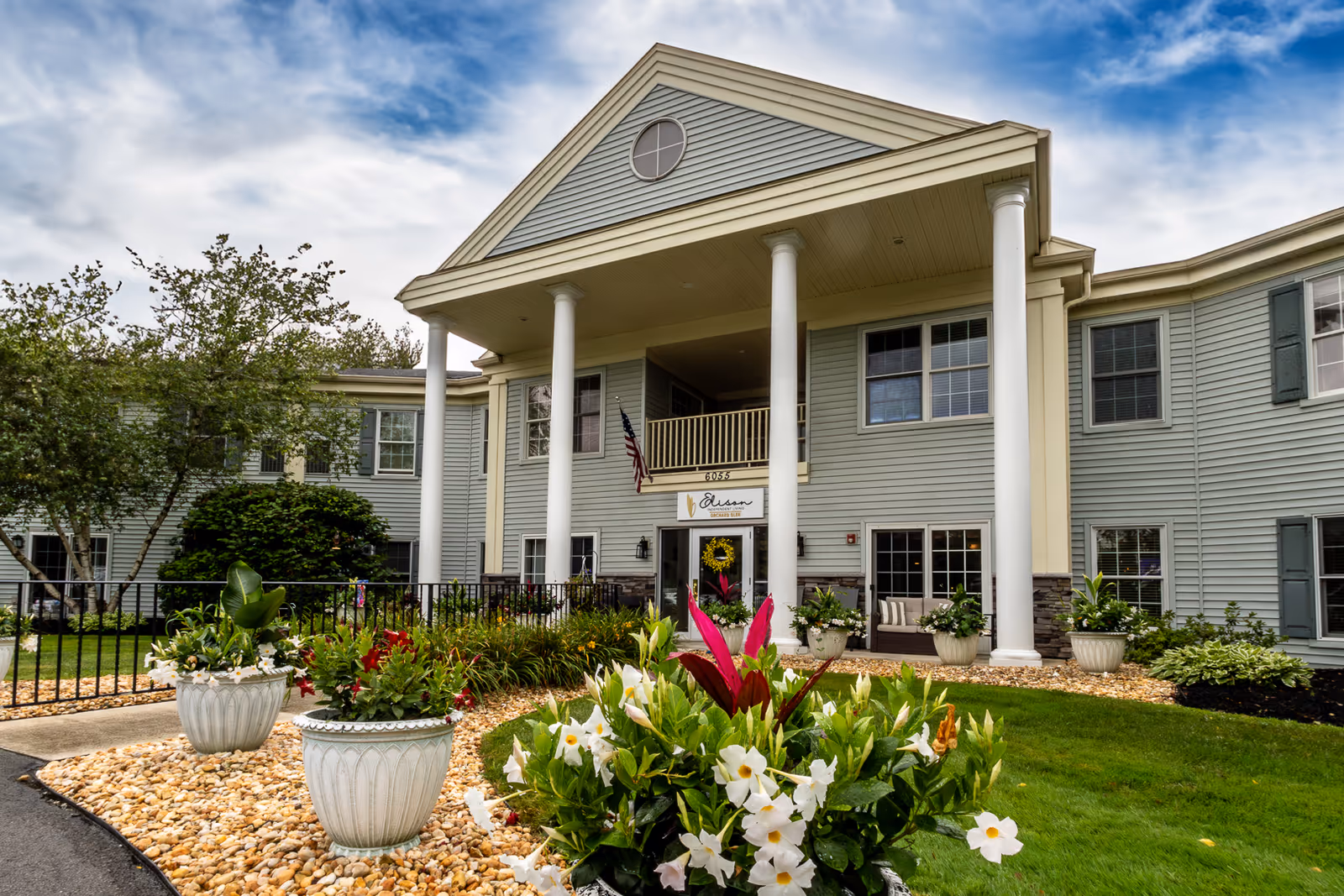 Front exterior view of Elison Independent Living of Orchard Glen, a two-story building with light gray siding, white columns, and a landscaped garden with flowers and shrubs in the foreground under a partly cloudy sky.