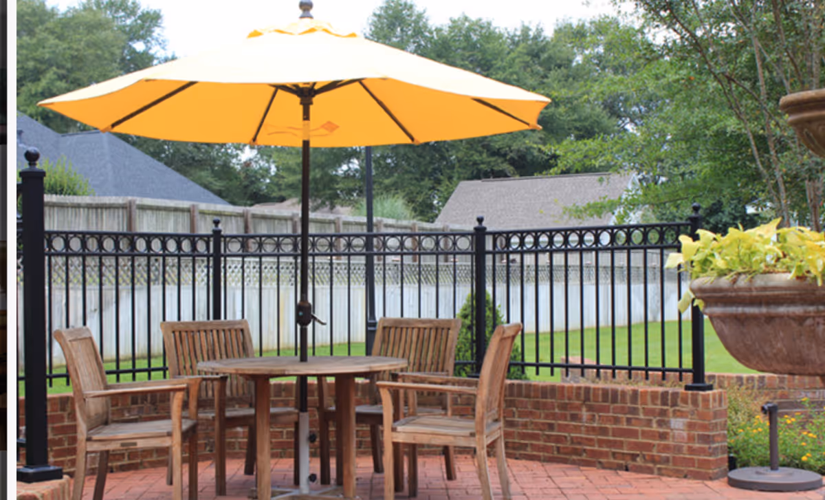 Outdoor patio area with a round wooden table and four wooden chairs under a large yellow umbrella. The patio is surrounded by a black metal fence and brick walls, with green grass and trees in the background.