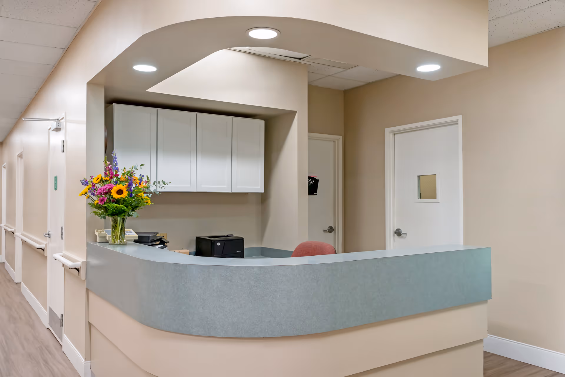 Reception desk area in a healthcare facility with a curved counter, a vase of colorful flowers, a printer, and white cabinets mounted on the beige wall behind. There are two closed white doors and ceiling lights illuminating the space.