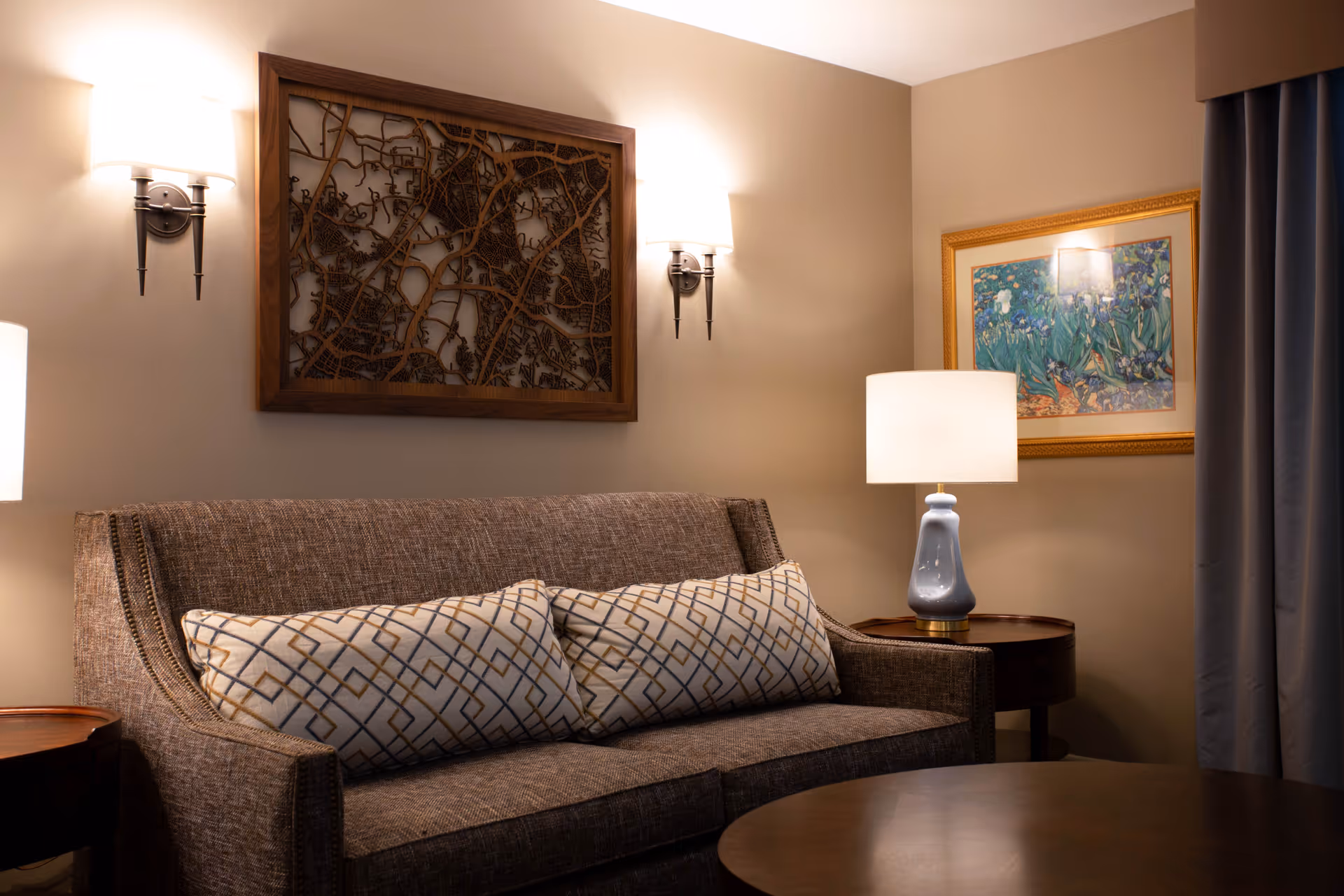 A cozy living room area with a brown upholstered sofa featuring two patterned cushions. Above the sofa is a decorative wooden wall art piece. On the right side, there is a round wooden side table with a blue ceramic lamp and a framed floral painting on the wall. Two wall sconces with white shades provide warm lighting, and a dark curtain is partially visible on the right.