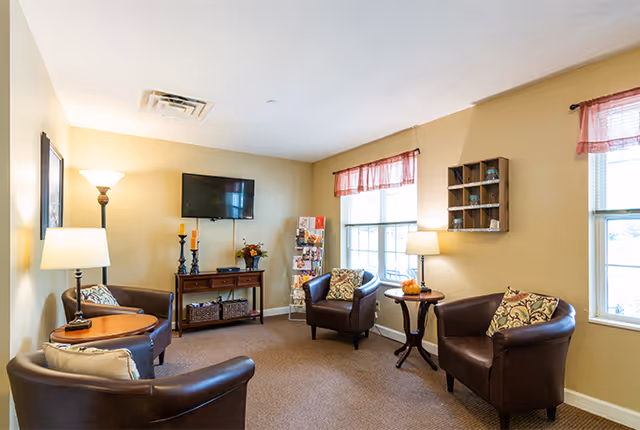 A cozy common sitting room with four leather armchairs arranged around side tables, a wall-mounted TV, lamps, and windows with valances.