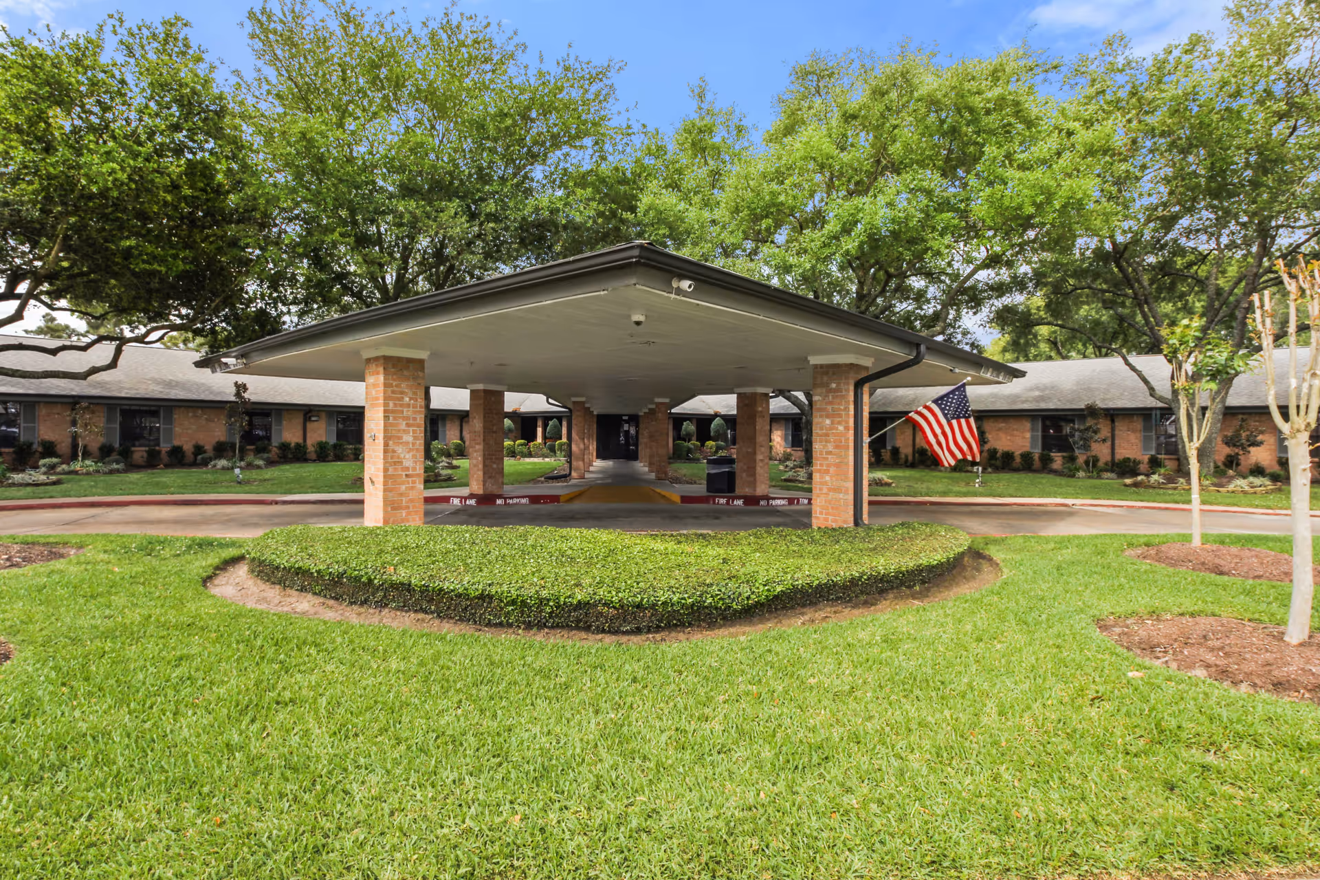 Front exterior view of Baywind Village Skilled Nursing and Rehabilitation showing a covered entrance supported by brick columns, a circular driveway, well-maintained green lawn, trees, and an American flag on the right side.