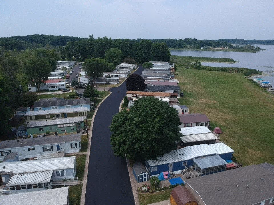 Aerial view of Lakeside Village 55+ Waterfront Community on Wabasis Lake showing rows of mobile homes along a paved road, surrounded by trees and green lawns, with the lake visible in the background.