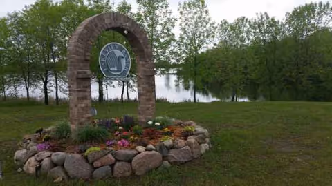 Brick arch sign surrounded by flowers and rocks, with trees and a lake behind.