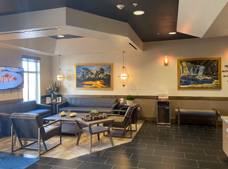 A cozy living room area in a senior living facility with modern furniture including a sofa, armchairs, and a coffee table. The room features two framed landscape paintings on the wall, pendant lights, a flat-screen TV mounted on the wall showing a food image, a water dispenser, and a bench. The floor is a combination of wood and dark tiles.