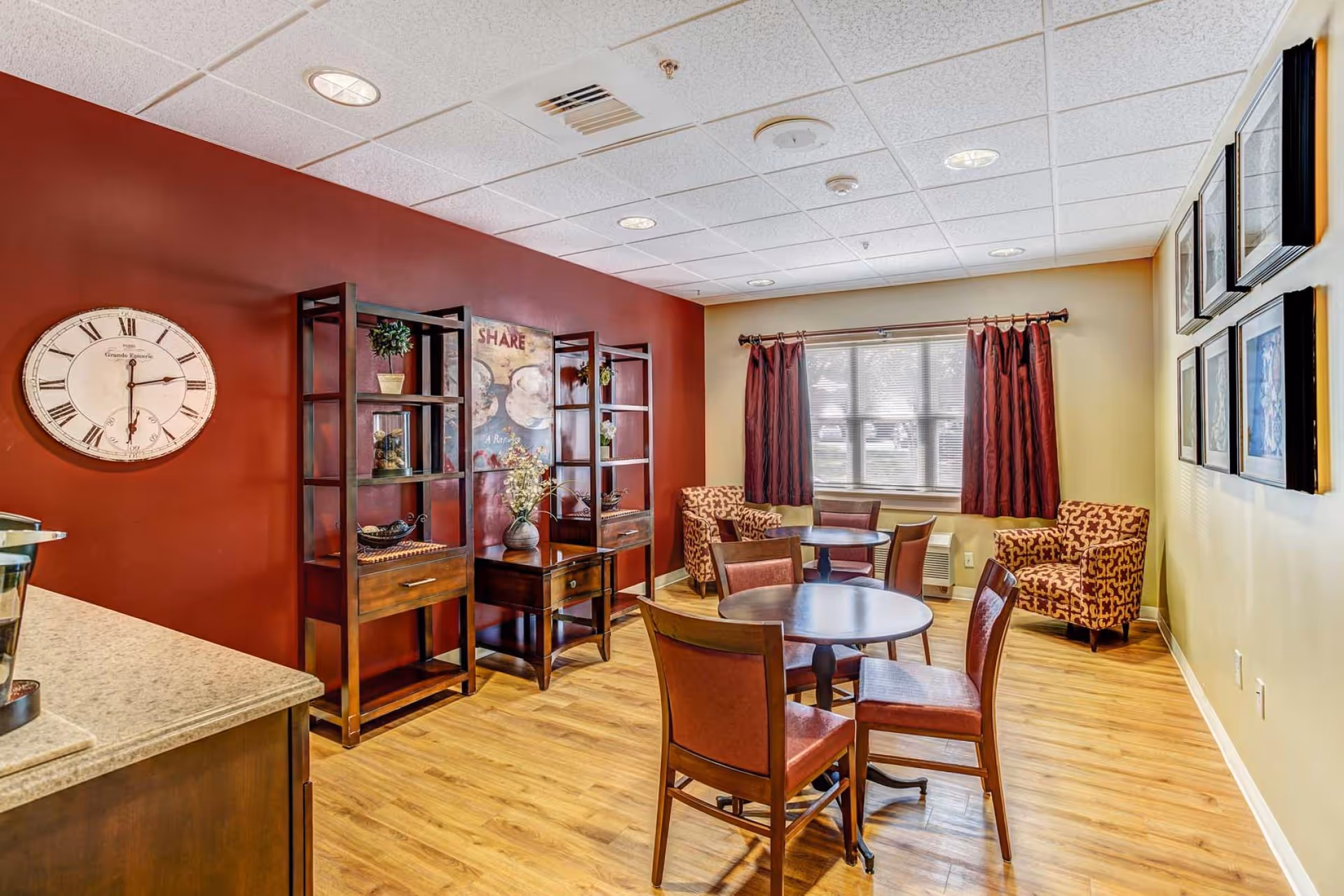 A cozy common area with wooden flooring, featuring two round tables each surrounded by four chairs with brown upholstery. Two patterned armchairs are placed near a window with red curtains. The walls are painted red and beige, decorated with framed pictures and a large round clock. Wooden shelving units with decorative items are against the red wall.