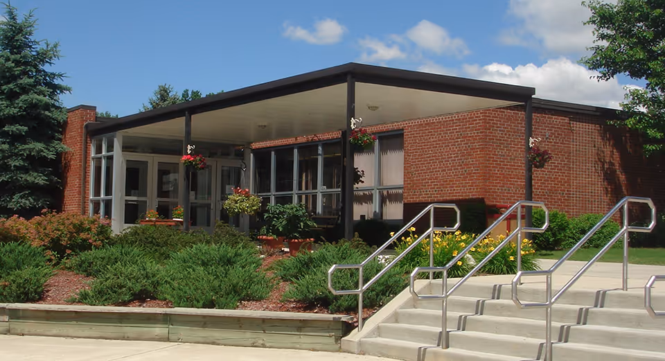 Front entrance of a single-story brick assisted living building with a covered entry, steps, metal handrails, and landscaped shrubs and hanging flower baskets.